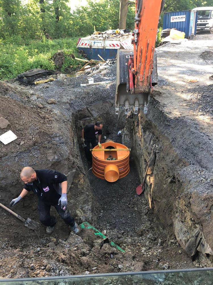 Zwei Männer arbeiten konzentriert in einer Baugrube mit einer großen orangefarbenen Röhre